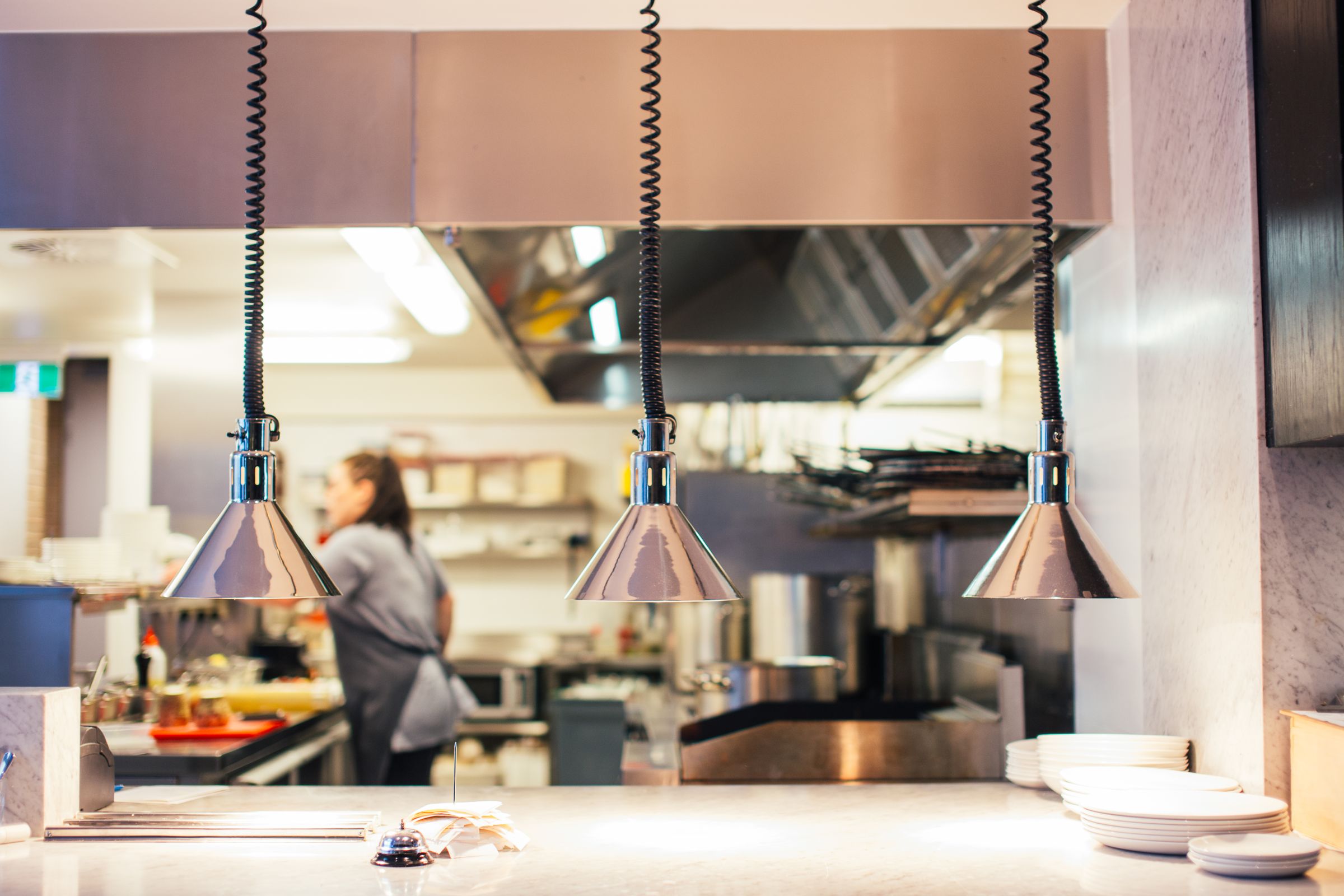 Three chrome heat lamps hang over a restaurant serving pass with stacked plates, while a cook works in the bright kitchen behind.