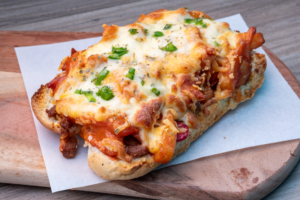 A toasted open sub topped with melted cheese, meat, sauce and chopped green peppers on paper on a wooden board.